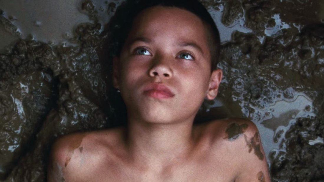 A young boy lays in the mud, looking up away from the camera. His chest is bare, and some of the mud has splashed onto his shoulders.