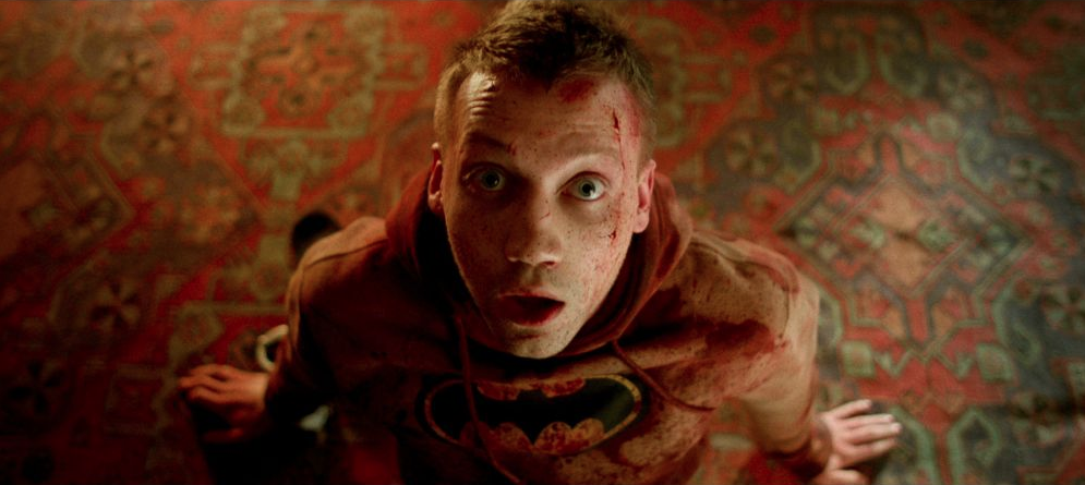 A young man in a batman hoodie is on the ground, framed by ornate carpeting, and is looking up at the camera. There is blood splattered across his face and the front of his hoodie and he is holding himself upright with his hands. He appears surprised.