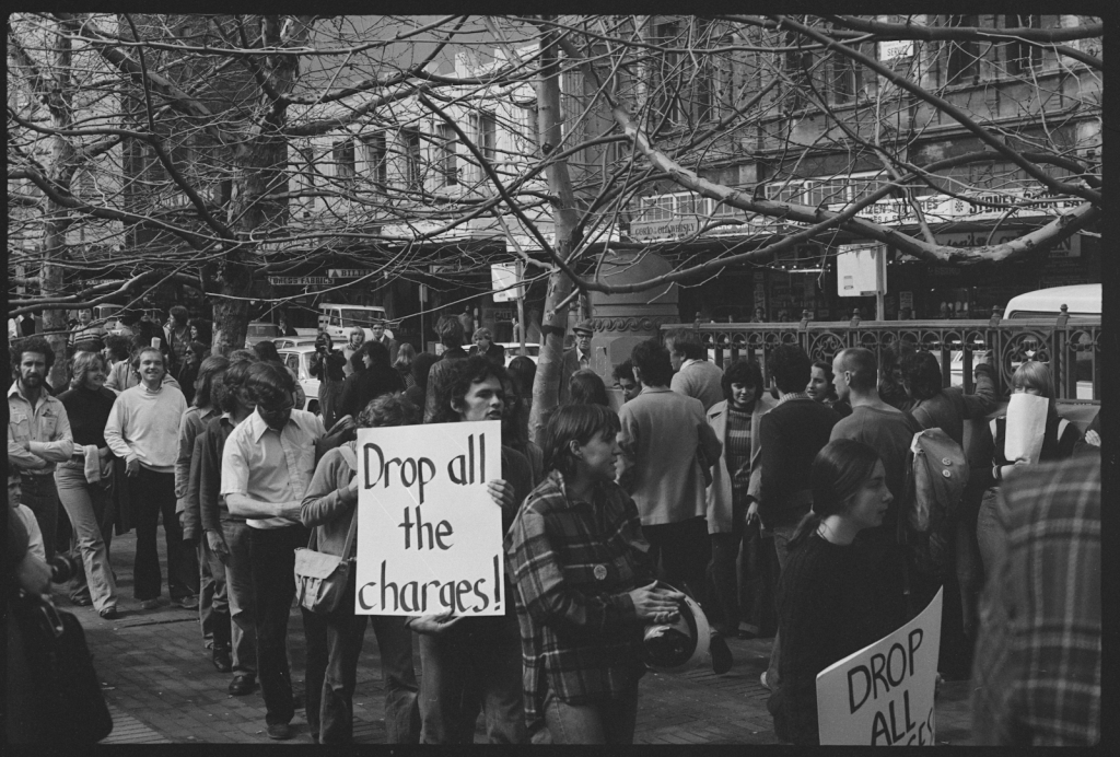 A Gay Liberation protest in New South Wales circa 1967. A man in the foreground is holding a white sign with black text that reads “Drop all the charges!