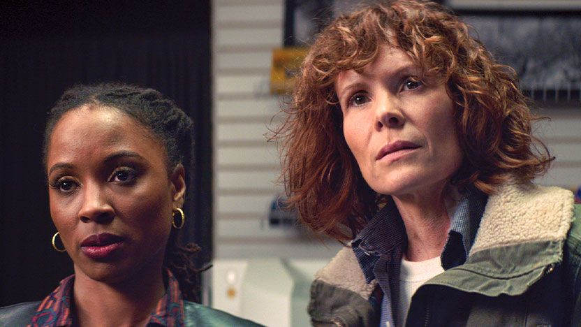 Two women look past the camera at someone, on the right is a red-haired White woman in "butch" clothing and next to her is a shorter Black woman with black hair that is pulled back. She is wearing earrings and a collared shirt.