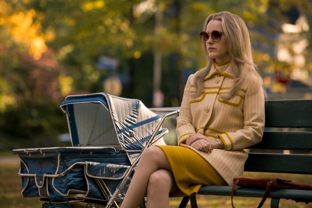 A blonde woman sitting alone on a bench at a park with a baby stroller right beside her.