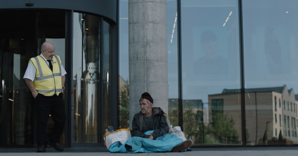 A middle-aged homeless man sits against a pillar in front of the large windows of an office building. To the side stands a security guard in high vis. From 'Pavement' 