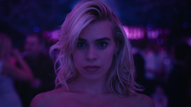 Image is from the TV Show 'I Hate Suzie'. A woman stares straight ahead as if she's focused on something. She is covered by purple lighting and seems to be at a nightclub.