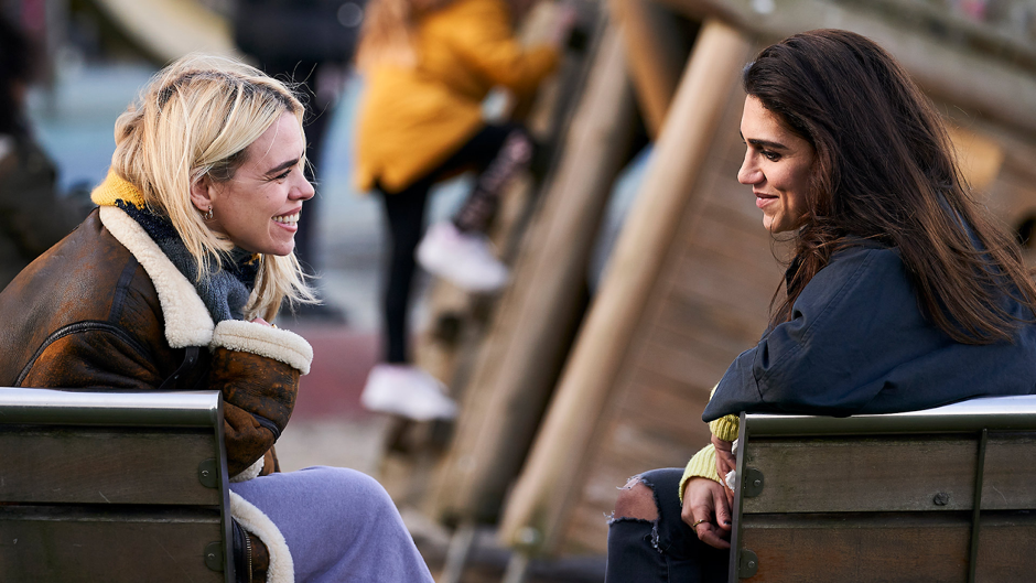 Suzie Pickles (Billie Piper) is sat on a park bench laughing with  Naomi Jones (Leila Farzad) who is sat on a park bench opposite her. 