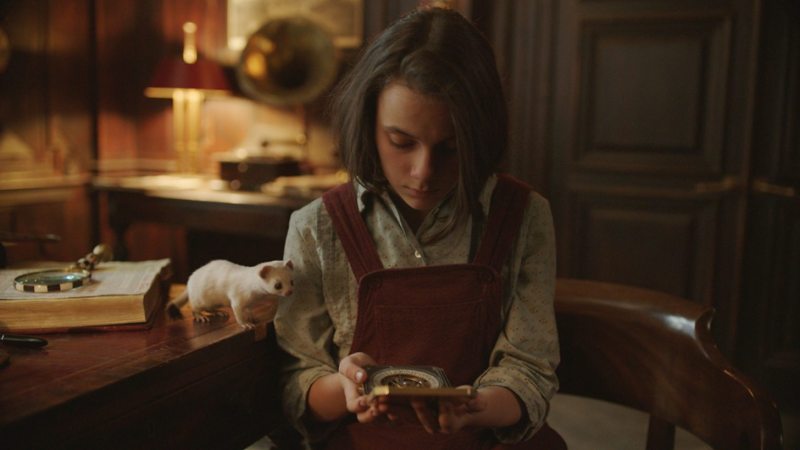 Image is from the TV Show 'His Dark Materials'. ‘Lyra Belacqua (Dafne Keen), a brown haired white girl wearing a red pinafore, studies a golden compass she holds in her hand. She sits in an academic office and her daemon Pan stands curiously on the desk next to her, also looking thoughtfully at the compass. He is in the form of a white ermine.