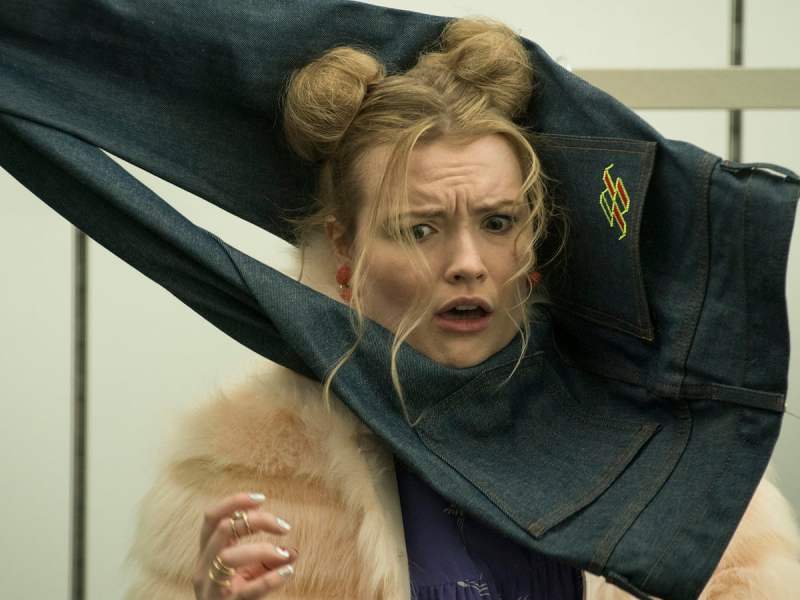 Image is from the film 'Slaxx' (2020). A young woman wearing a fur coat and space buns is being strangled by a pair of jeans.