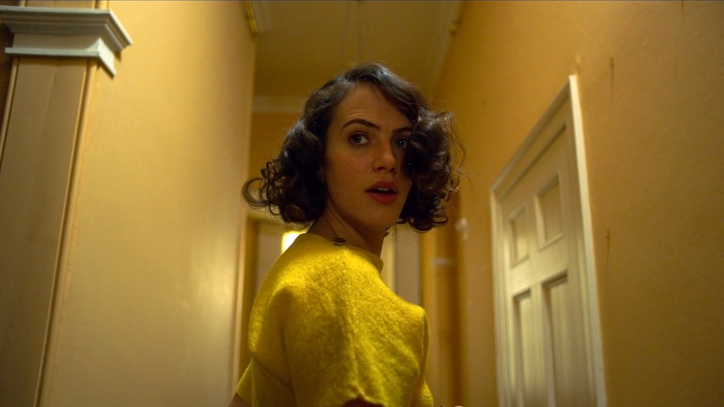 Marianne (Jessica Rose Findlay), dressed in a yellow top, is turned in her hallway looking back at something unseen