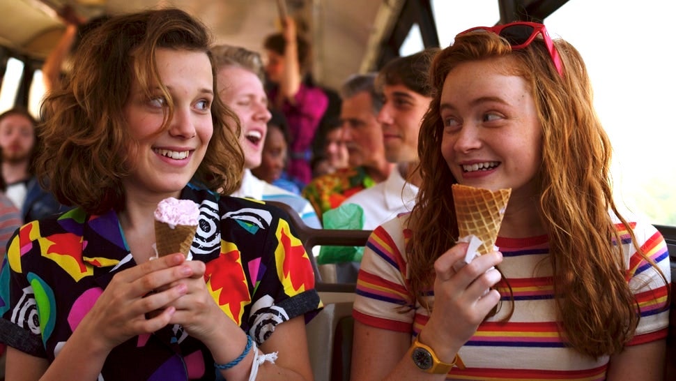 Image is from the TV Series 'Stranger Things'. Two girls sit beside one another on a bus full of people. Both are wearing brightly coloured, patterned tops and are holding ice cream.