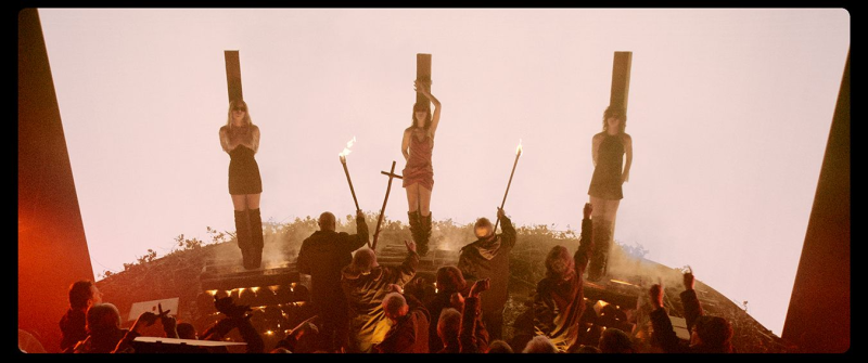 Three women who are tied to wooden stakes while wearing revealing outfits. They are seen from a distance, with figures surrounding them and brandishing torches.