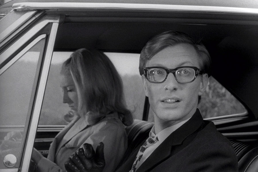 A black and white image of a man and woman in a car. The woman is seen from the side, looking away, while the man wears glasses and appears interested in what's outside the window.