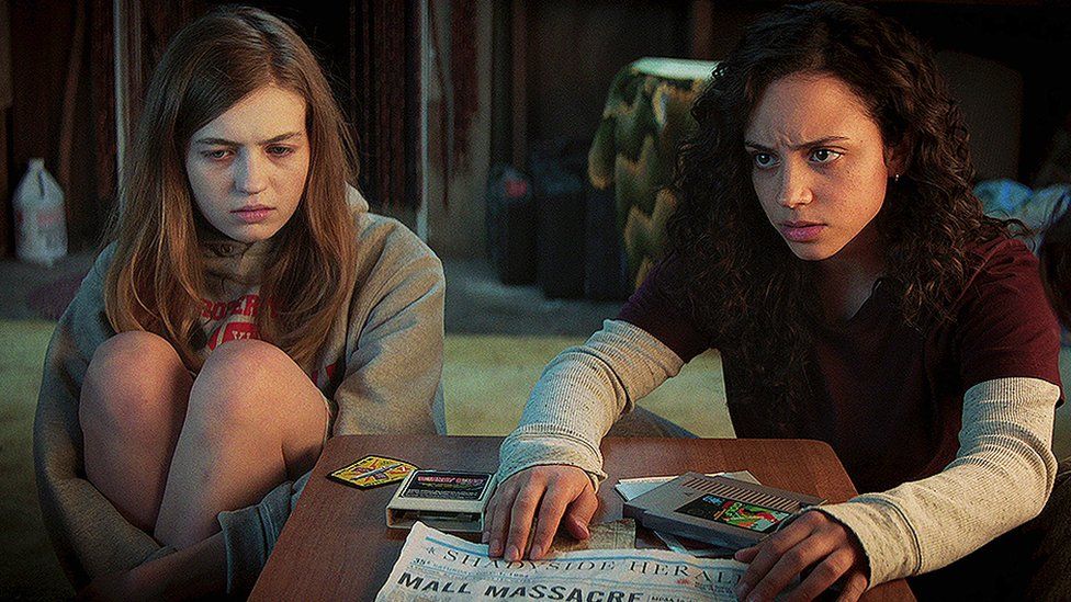 Olivia Scott Welch as Samantha and Kiana Madeira as Deena in 'Fear Street Part One': 1994, looking pensive over a coffee table with a newspaper that says MALL MASSACRE.