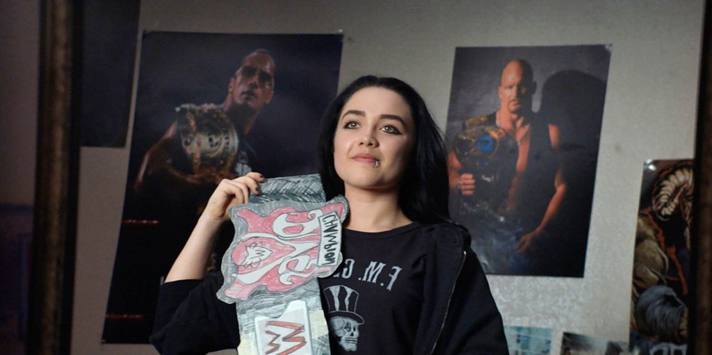 Saraya (Pugh) stands in front of posters of pro wrestlers in her bedroom, looking in the mirror. She has black hair with dark makeup and clothes, and looks proud as she holds up a paper WWE Divas Championship Belt.