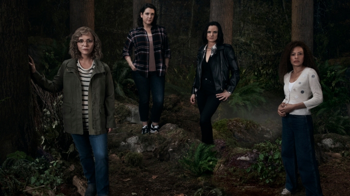 A promotional photo for 'Yellowjackets' that shows four women standing in a forest. They are dressed in clothes that range from casual to formal, with varying poses.