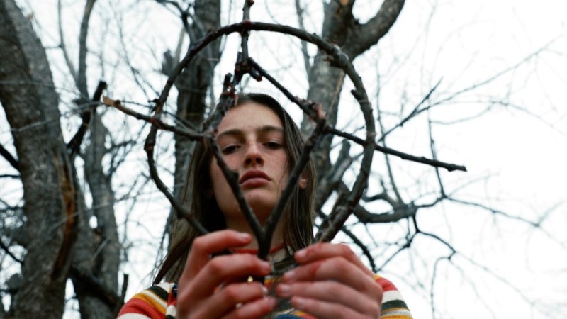 Izzy (Zelda Adams) fashions a pentagram out of twigs. She is looking down at it and wearing a colourful striped shirt.