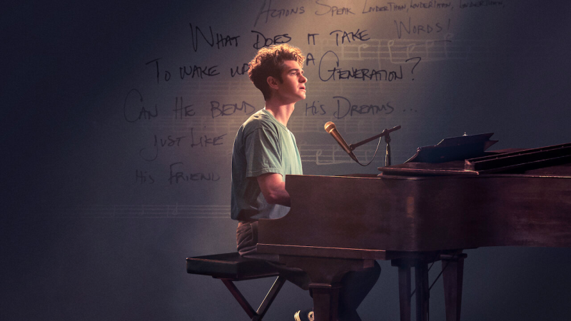 Andrew Garfield as Jonathan Larson in 'Tick, Tick... BOOM!'. He sits at a piano while singing with a pensive expression on his face, wearing a green t-shirt. Words and a music score can be seen in the background behind him.