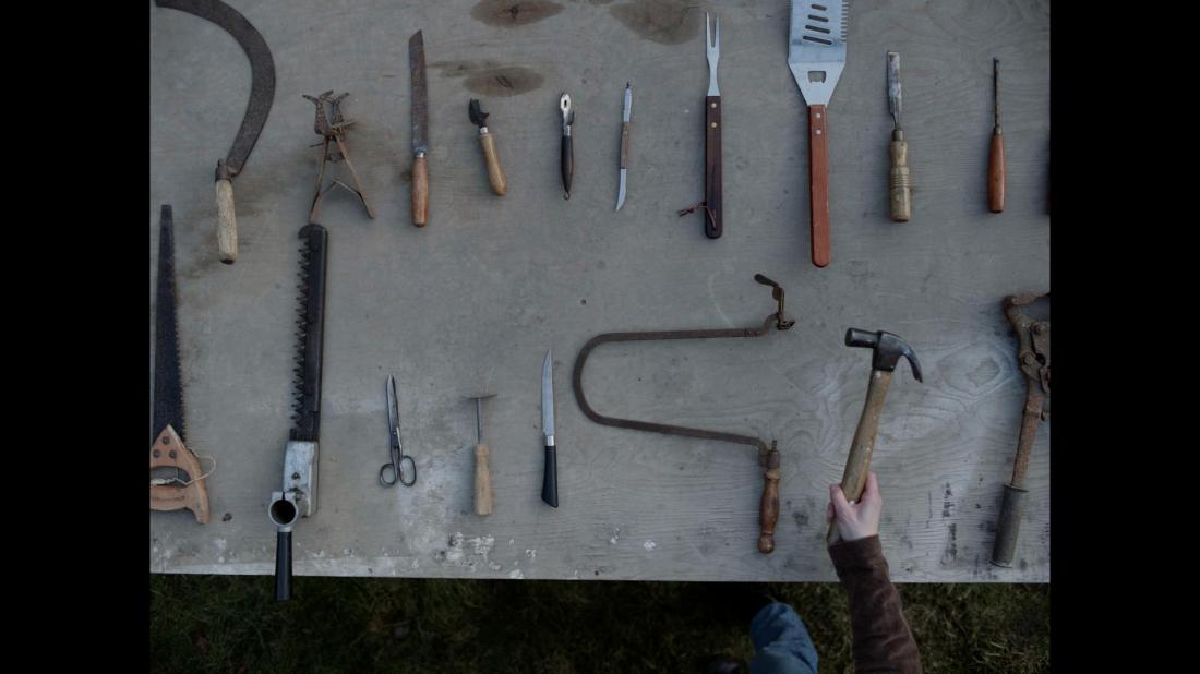 Still from The Retreat taken from above showing a bench filled with various instruments and tools including a hammer, scissors, and various kinds of knives. A person is seen reaching out into the frame, holding a firm hand on the hammer as if contemplating using it out of the big selection available.