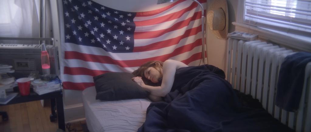 Still from Undress Me showing the female protagonist waking up alone after her sexual encounter with a frat boy. Behind the bed is the flag of the United States of America hanging, as well as a cowboy hat. On a nightstand is a stereo, lava lamp and a red solo cup visible. 