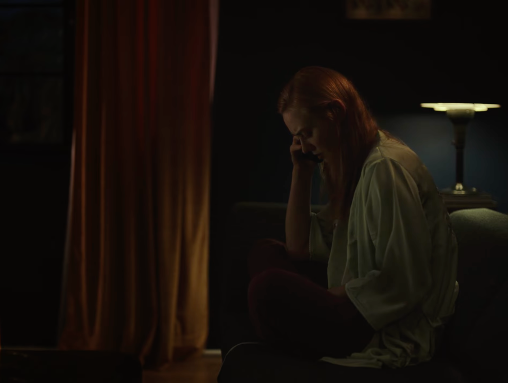 Still from Willing to Go There portraying the female protagonist sitting inside of her apartment and talking on her phone with the strange man. It's evening and the sole light source comes from a desk lamp diagonally behind her. She's sitting with her legs up on the sofa, crossed, and she's looking down at her lap with a concerned facial expression.