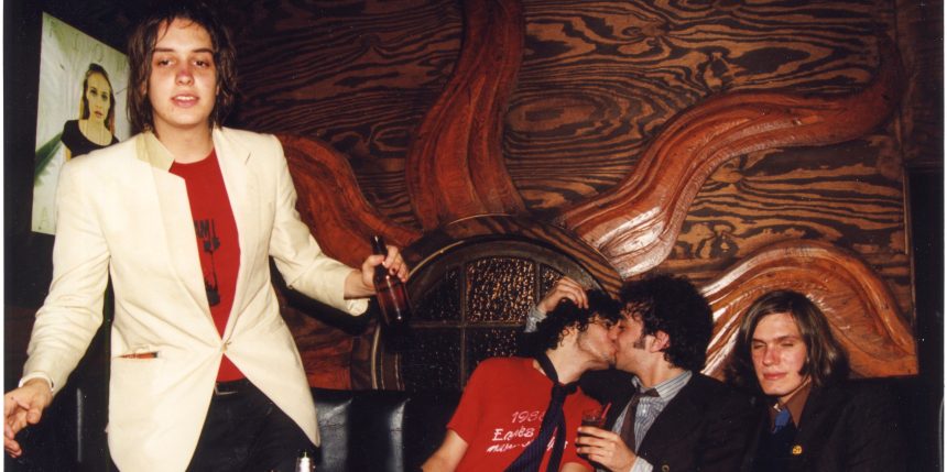 The Strokes are positioned in a dark-wood room. Julian Casablancas stands with a beer bottle in his hand, while the other bandmates sit on a couch. Two of them are kissing. There is a Fiona Apple poster in the top left corner.