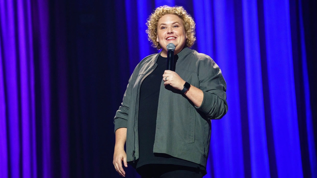 Still from Good Fortune showing its performer Fortune Feimster from the hips and up whilst standing on stage, smiling big with a microphone close to her mouth looking out towards her crowd.
