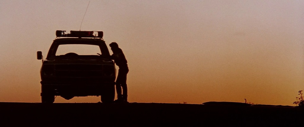 Still showing the ending, with Jim Halsey standing by the side of a cop car against the orange-hued sunset. It's a moment of solitude and reflection. 