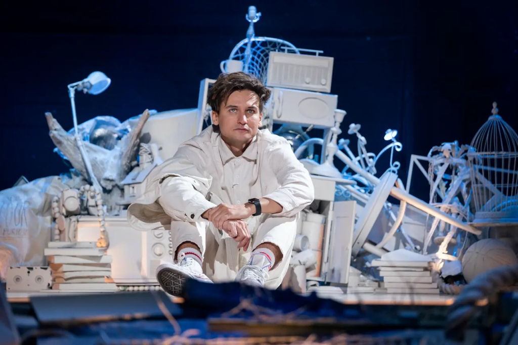 Still showing Moses sitting on stage with his arms around his knees. He is wearing all white and is looking up towards something out of frame. Behind him are various objects and junk (including a birdcage, a chair, a VHS tape, books, and a candle holder) shown together in a pile, all painted in white. Moses has a neutral facial expression, almost something sad about it in his eyes.
