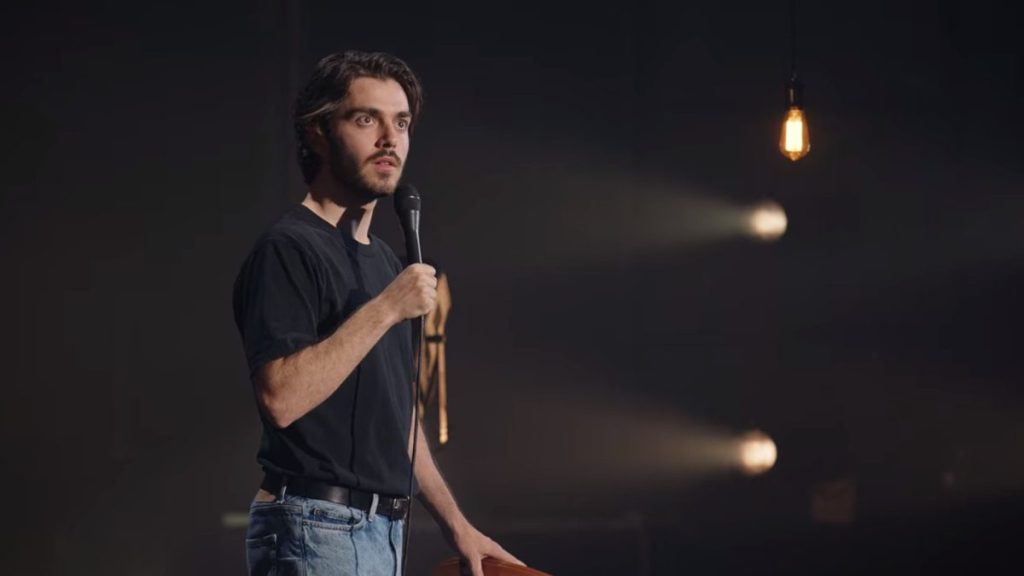 Still showing Panayotis from the hips and up with a dark and, besides some spotlights and a hanging lightbulb, a barely lit background. He is wearing casual clothes — light blue jeans, black belt, and a black t-shirt — and is looking out over the crowd with a surprised facial expression whilst his microphone is close to his mouth.