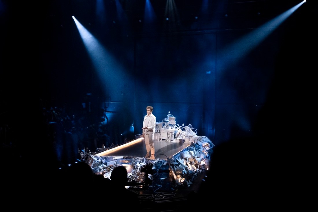 Still showing Moses Storm (Trash White) from a distance, as he is standing on his stage that is surrounded by various objects and junk painted white. He is wearing all white, and the spotlights highlight both him and his stage. Image courtesy of HBO Max.