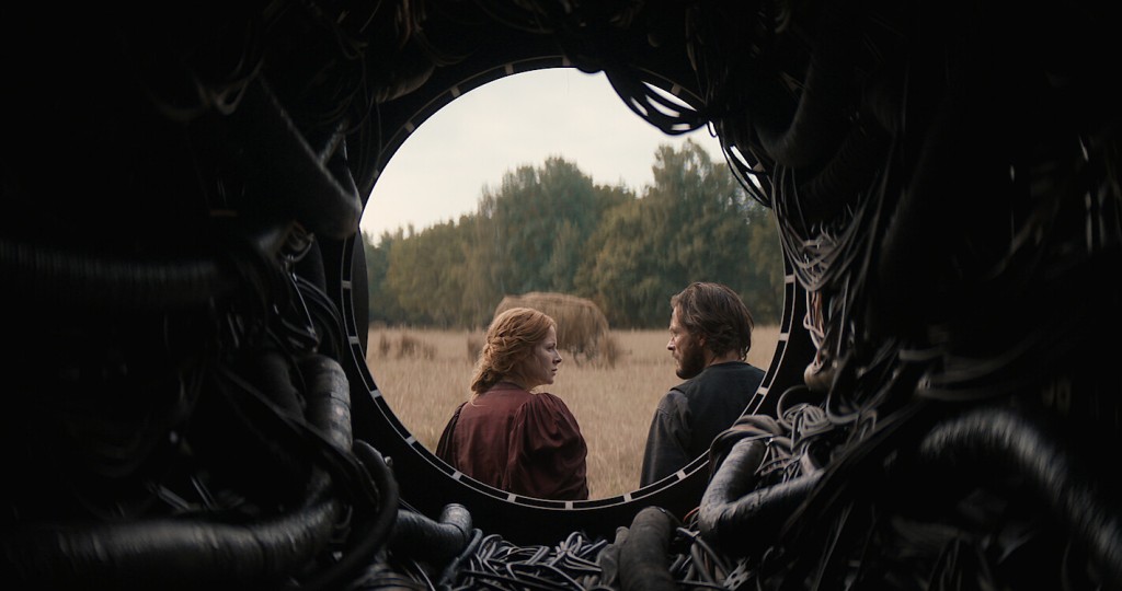 Maura and Eyk climbing into a memory. They are positioned within a circle as the shot is taken from inside the cylindrical pathway they have just exited. Within the pathway, visible cables of various sizes are seen. Maura and Eyk are looking at each other with serious facial expressions. In front of them is a hay field with lightly browning grass as well as a green forest in the distance and a bright, almost white, coloured sky. 
