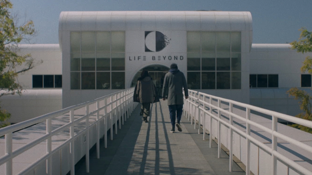 A still of Rose (Katie Parker) and Teddy (Rahul Kohli) from 'Next Exit'. They are seen outside walking down a ramp and into a modern white building emblazoned with the text "LIFE BEYOND" and a black and white semicircle logo.