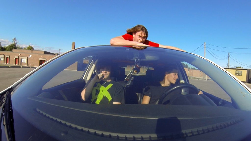 The still is positioned from the point of view of a car's hood. Inside it are two teenagers sitting in the driver's and passenger's seat, respectively. On top of the car is Justin seen clinging on as hard as he can as he is strapped to the roof of the car. They are in an empty parking lot.