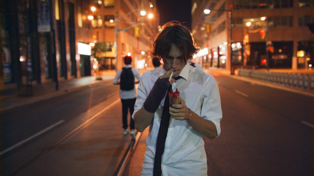 It’s nighttime out on the streets of the big city. Justin is seen centered in the middle of the still, with a bandage on his right hand, whilst smoking a cigarette. Cigarette in mouth, he is about to soon light up the fireworks he is holding in his left hand by using the lit cigarette. Behind him are the lights of the city shining out of focus, along with his friends standing with their backs against the camera further down the street.
