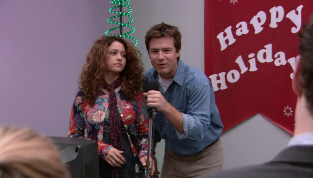 Maeby and Michael seen on stage during the first Christmas party singing karaoke together. In the background, a green neon decoration in the shape of a tree is seen as well as a sign saying "Happy Holidays." 