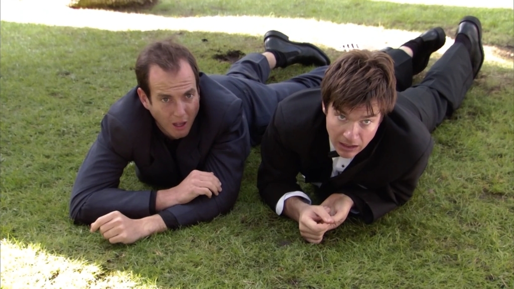 Gob and Michael are both wearing formal clothing as they are fighting on the ground outside of the courthouse.  They are in the green grass looking up towards someone outside of frame. 