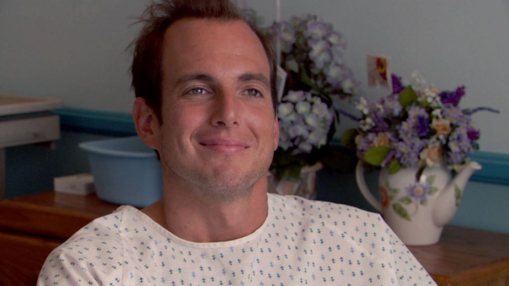 Close up of Gob wearing a hospital gown and staring dreamily into the distance outside of the frame. Behind him are vases of flowers seen. 