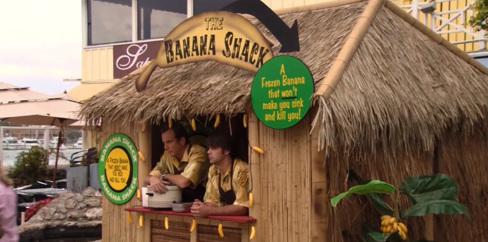 Gob and his son Steve seen standing inside their competitive banana stand. Their business is called "The Banana Shack" and their tagline is seen being "A Frozen Banana that won't make you sick and kill you." 