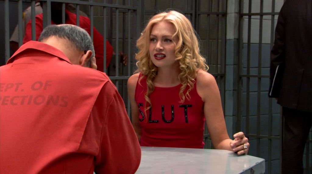 Lindsay is in prison, seen sitting in front of her father George Sr. who is only seen from behind in this still. He is wearing an orange jumpsuit and Lindsay is wearing a red tank top with black bedazzled letters spelling out the word "SLUT." She has a distressed facial expression. 