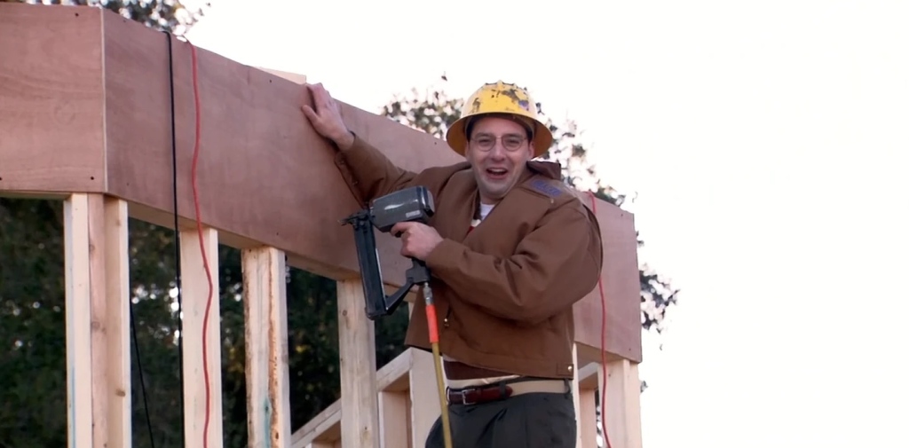 Buster working on the construction site, wearing a yellow security helmet and smiling big as he is looking towards the camera. He is holding a power tool and is standing on a ladder (not shown in the frame). 
