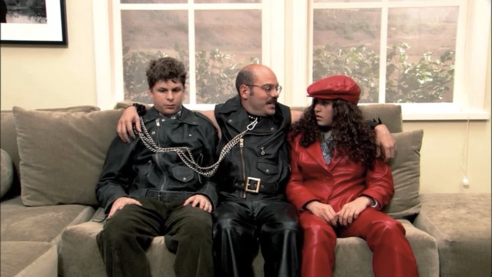 George Michael, Tobias, and Maeby are sitting on the sofa in the model home. They are all wearing leather outfits, with George Michael and Tobias wearing black while Maeby stands out in red. George Michael looks uncomfortable, Maeby looks confused and Tobias looks pleased as if he is a part of their friend group. 