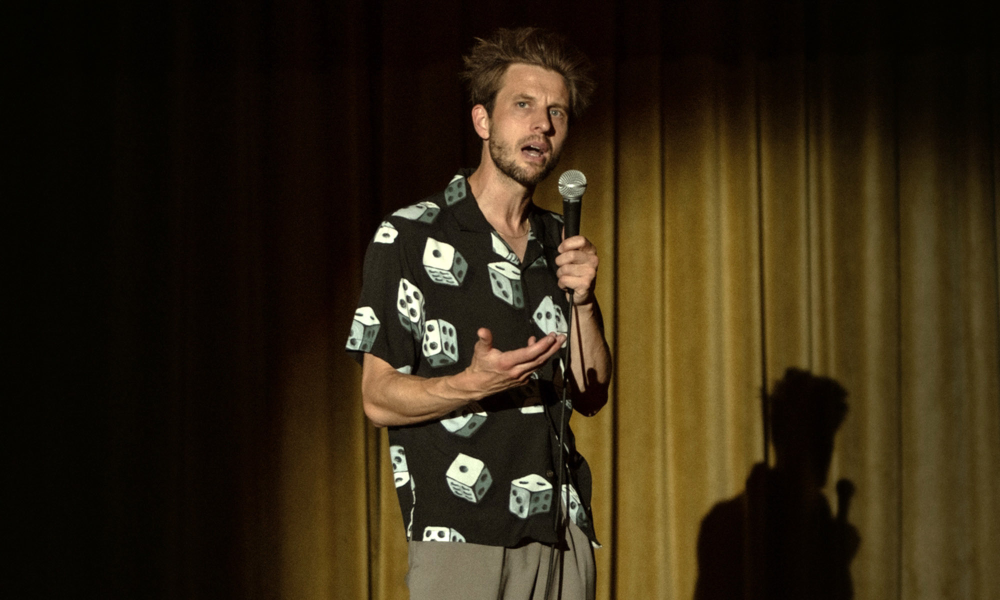 David (Anders Danielsen Lie) seen performing his stand-up set. There’s a spotlight on him, highlighting David and creating shadows around him along with a shadow of himself behind him. He’s in the middle of talking, one hand holding a microphone and the other gesticulating. He is wearing a black buttoned shirt with short sleeves and a printed pattern of white dices with black dots on them. 