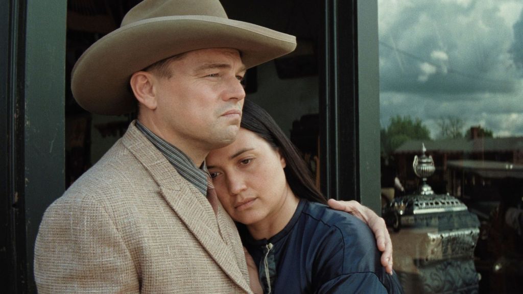 A still of Ernest Burkhart (Leonardo DiCaprio) and his wife Molly (Lily Gladstone) from 'Killers of the Flower Moon'. Ernest has an arm around Molly, who is learning into him, and they stand against a doorframe.