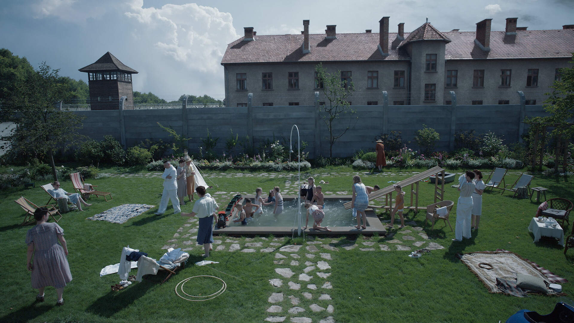 A still from the film 'The Zone of Interest'. People are shown outside lounging in chairs and swimming, among other outdoor activities. A fence