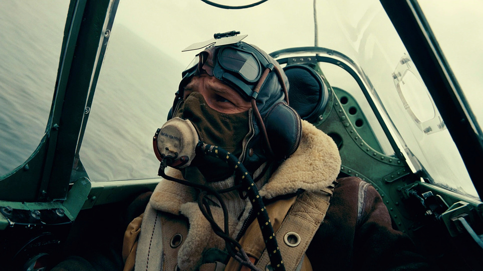 A still from the film 'Dunkirk' of a fighter pilot in a plane wearing a mask and bomber jacket.